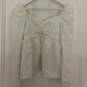 Vit blus från BikBok - Säljer en elegant vit blus från BikBok med subtilt mönster och långa ärmar. Blusen har en smickrande passform med rynkade detaljer vid axlarna och en lätt volang nedtill. Perfekt för en stilren look.
