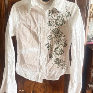 Vit och beige skjorta från Free Soul Italia - Snygg vit och beige skjorta från Free Soul Italia med unikt blommönster i grönt. Skjortan har långa ärmar och knappar framtill. Perfekt för dig som vill ha något stilrent med en twist. !Obs! Det saknas en knapp nertill se bild men går lätt att sy på en knapp eller ha den som den är.