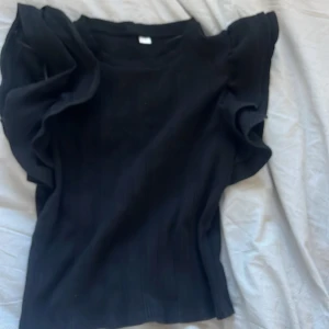 Svart topp med volangdetaljer - Snygg svart topp från Gina Tricot med volangdetaljer på ärmarna. Toppen har en ribbad struktur och en elegant design som passar perfekt för olika tillfällen.