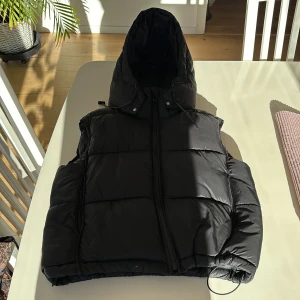 Svart dunväst från Zara - Säljer en svart dunväst från Zara med dragkedja och huva. Perfekt för kyligare dagar och ger en stilren look. Västen har en normal passform och är lätt att matcha med olika outfits.