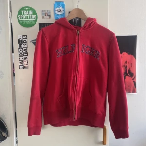Röd hoodie från Tommy Hilfiger - Säljer en snygg röd hoodie från Tommy Hilfiger med dragkedja och huva. Den har ett broderat Hilfiger-logotyp på framsidan och ribbade muddar. Perfekt för en avslappnad stil. Passar xs/s/m