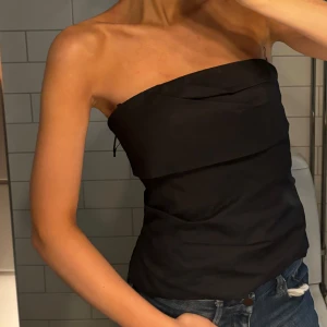 Svart axelbandslös tubtopp - Snygg svart axelbandslös tubtopp i ett stilrent och enkelt utförande. Perfekt för en kväll ute eller en avslappnad dag. Passar bra med jeans eller kjol för en trendig look.
