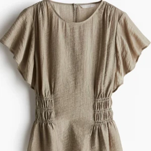 blus - Snygg beige blus med korta ärmar och elastisk midja som ger en smickrande passform. Tillverkad i ett lätt och luftigt material, perfekt för varmare dagar.