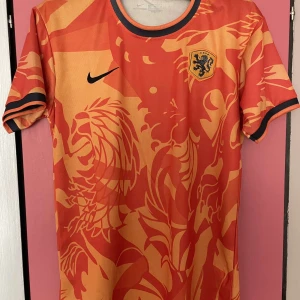 Orange fotbollströja från Nike - Snygg orange fotbollströja från Nike med ett lejonmönster och svarta detaljer. Tröjan har korta ärmar och ett runt halsband. Perfekt för fotbollsfans som vill sticka ut på planen eller läktaren.