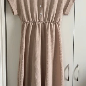 Beige klänning med knappar - Säljer en stilren beige klänning med korta ärmar och knappar framtill. Klänningen har en elastisk midja som ger en fin passform. Perfekt för en avslappnad look.