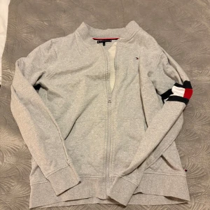 Grå kofta från Tommy Hilfiger - STORLEK : 176 = Som XS                                                      Säljer en stilren grå kofta från Tommy Hilfiger med dragkedja och ribbade muddar. Den har en liten logotyp på bröstet och en färgglad detalj på ärmen. Perfekt för en avslappnad look.