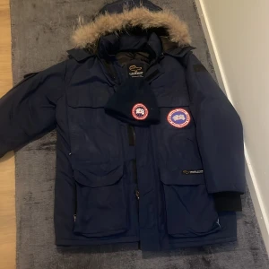 Blå parkajacka från Canada Goose - Säljer en blå parkajacka från Canada Goose med pälsdetaljer på huvan. Jackan har flera praktiska fickor och en dragkedja framtill. Perfekt för kalla vinterdagar. 🧥❄️