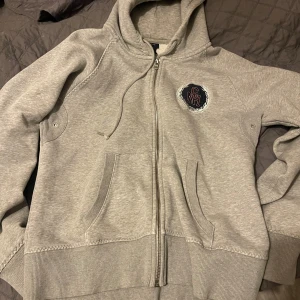 Grå hoodie med dragkedja - Säljer en grå hoodie med dragkedja och en broderad logga på bröstet. Den har en justerbar huva och praktiska fickor framtill. Perfekt för en avslappnad stil.