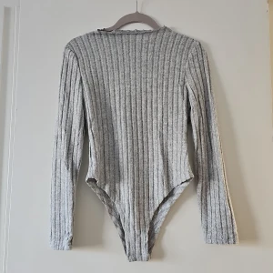 Grå ribbad bodysuit från Shein - Säljer en stilren grå ribbad bodysuit från Shein i storlek petite S. Den har långa ärmar och är tillverkad i en mjuk polyesterblandning. Perfekt för lager-på-lager eller att bära som den är.