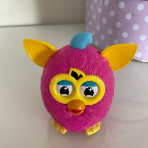 Furby leksaksfigur från Hasbro - En färgglad Furby leksaksfigur från Hasbro, tillverkad för McDonald's 2014. Den är rosa med gula öron och blå detaljer. Perfekt för samlare eller som en rolig leksak för barn.Viftar på öronen när man kör den  Ca 9 cm hög.kan möta er i Stockholm utanför Åhléns 
