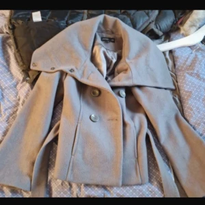 Beige kappa från Sara Kelly - Supersnygg grå kappa från Sara Kelly❤️ Det går att ha kragen på flera olika sätt och midjebandet är avtagbart. Den är alltså anpassbar efter ens egna preferenser!😍 Notera att färgen som sagt är grå och inte beige som kameran uppfattar ibland 