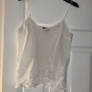 Vit linne med spetsdetaljer - Säljer ett vitt linne med tunna axelband och v-ringning. Linnet har spetsdetaljer och broderier längs nederkanten. 