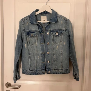 Jeansjacka från River Island - Snygg jeansjacka från River Island i ljusblå denim med slitna detaljer. Jackan har knappar framtill och två bröstfickor med knappstängning. Perfekt för en cool och avslappnad stil. Passar xs oxå