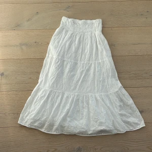 Vit långkjol från Gina Tricot - Charmig vit långkjol från Gina Tricot med broderade blommor och elastisk midja. Perfekt för en somrig look med sin lätta och luftiga design. Kjolen har en romantisk känsla med sina fina detaljer och passar utmärkt för varma dagar.