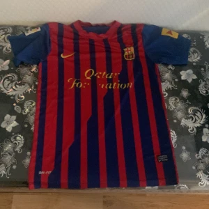 Blårandig FC Barcelona fotbollströja - Säljer en klassisk FC Barcelona fotbollströja från Nike med röda och blå ränder. Tröjan har korta ärmar och klubbens emblem på bröstet. Perfekt för alla Barça-fans!