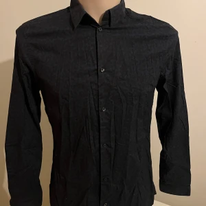 Svart och blå mönstrad skjorta från H&M - Snygg svart och blå mönstrad skjorta från H&M i premium bomull. Skjortan har långa ärmar och knappar framtill. Perfekt för en stilren look.