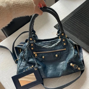 Blå Balenciaga väska - Kollar intresset på min älskade Balenciaga väska i skinn med guldfärgade detaljer och dragkedja. Väskan har en praktisk spegel och flera fack för enkel organisering. Perfekt för att bära med sig det viktigaste med stil. Sista två bilderna visar hur stor den är❤️ köptes här på Plick för 6500kr!