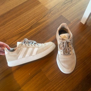 Beige sneakers från Adidas - Säljer ett par stilrena beige sneakers från Adidas med klassiska tre ränder i vitt på sidan. Skorna har snörning och en bekväm sula, perfekt för vardagsbruk.