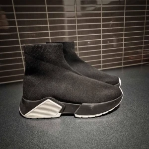 Svarta sneakers med vit sula - Snygga svarta sneakers med en elastisk stickad överdel och en kontrasterande vit sula. Skorna har en modern design med en högre ankel och en bekväm passform.