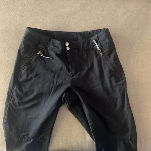 Svarta byxor med dragkedja och knappar - Säljer ett par svarta byxor i syntetmaterial. De har en snygg och praktisk design med dragkedjor på fickorna och knappar i midjan. Byxorna har en justerbar snörning vid bensluten för en perfekt passform. Perfekta för höst och vår!