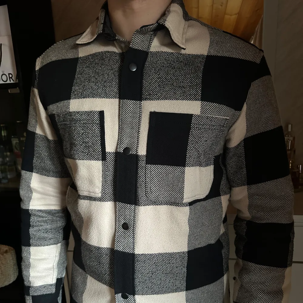 Säljer en snygg svart och vit rutig overshirt. Den har en klassisk krage och knappar framtill. Två stora bröstfickor ger den en cool och praktisk look. Perfekt för lager-på-lager-stil under höst och vår. Passar både till vardags och lite mer uppklädda tillfällen.. Jackor.