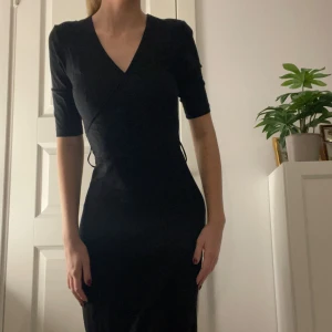 Svart knälång omlottklänning - Säljer en elegant stretchig svart omlottklänning med v-ringning. Klänningen har trekvartsärmar och en smickrande passform som passar perfekt för både fest och vardag. Den är gjord i ett mjukt och bekvämt material som känns skönt mot huden. Passar S