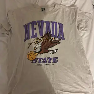 Säljer en beige t-shirt från FSBN med ett coolt tryck av en örn och texten 'Nevada State Athletics Allstar Champions 1985'. T-shirten är kortärmad och perfekt för en avslappnad stil. Passar bra till vardags eller för en sportig look! Baggy style 