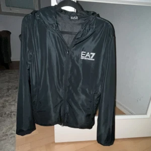Svart vindjacka från EA7 - Säljer en snygg svart vindjacka från EA7. Den har en dragkedja framtill och en huva för extra skydd mot väder och vind. Perfekt för vår och höst! Jackan är i ny skick.