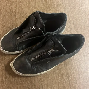 Svarta sneakers med dragkedja - Säljer ett par svarta sneakers från Arigato. Skorna är i skinn och har en vit sula som ger en snygg kontrast. De är lite slitna men fortfarande i ok skick, inte jätte bra. Strl 41 men passar 42. Skulle säga skick 5/6-10