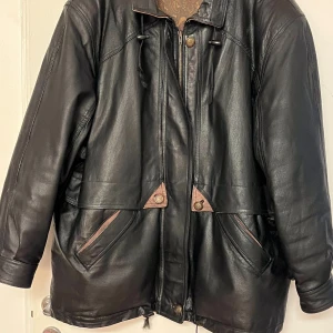 Vintage Svart skinnjacka med mönstrat foder - Vintage Snygg svart skinnjacka med dragkedja och knappar. Jackan har ett unikt mönstrat foder och praktiska fickor framtill. Perfekt för höst och vår.