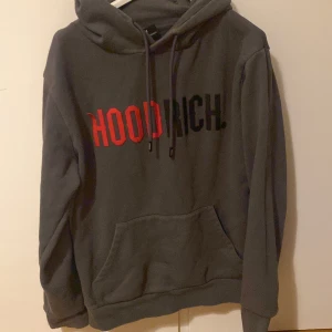 Grå hoodie från Hoodrich - Säljer en grå hoodie från Hoodrich med texten 'HOODRICH' i rött och svart på bröstet. Tröjan har en klassisk känguruficka och justerbar huva med dragsko. Perfekt för en avslappnad stil.