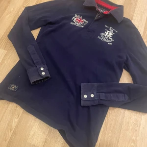 Mörkblå pikétröja från Morris - Snygg mörkblå pikétröja från Morris med broderade detaljer och brittisk flagga på bröstet. Tröjan har långa ärmar och en klassisk krage med knappar. Perfekt för en stilren look.                            -Snålt använd                                                                 -Köpt på NK