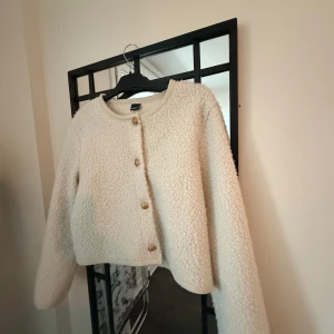 Beige teddyjacka med knappar - Mysig beige teddyjacka med rund hals och knappar framtill. Perfekt för kyliga dagar med sin fluffiga textur och stilrena design.