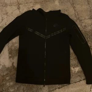 Nike tech fleece - Snygg svart jacka från Nike med dragkedja och diskret logga på bröstet. Koftan har långa ärmar och en stilren design, perfekt för en sportig look. Köpte den för 1200 