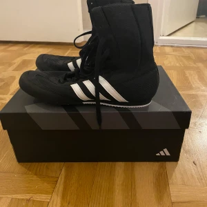 Svarta Box Hog 2 skor från Adidas - Säljer ett par svarta Box Hog 2 skor från Adidas med vita ränder. Skorna har hög snörning och är perfekta för boxningsträning. De är designade för att ge bra stöd och grepp. Bara använda några gånger och kommer med kartongen!