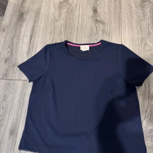 Mörkblå topp från Holly & Whyte - Snygg mörkblå topp från Holly & Whyte med korta ärmar och rund halsringning. Perfekt för en stilren look med en subtil rosa detalj på insidan av kragen.