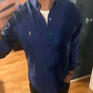  Ralph Lauren Skjorta - Snygg blå skjorta från Ralph Lauren med klassisk krage och broderad logotyp på bröstet. Perfekt för en stilren look. Skjortan har långa ärmar och knappar framtill.