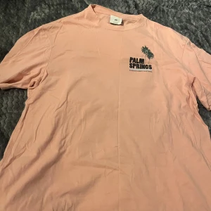 Rosa t-shirt från H&M - Säljer en rosa t-shirt från H&M med ett coolt Palm Springs-tryck. T-shirten har en avslappnad passform och är perfekt för en somrig look. Den är kortärmad och gjord i mjukt bomullsmaterial. 