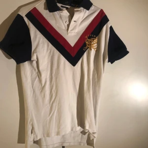 Vit pikétröja från Ralph Lauren - Säljer en stilren vit pikétröja från Ralph Lauren med marinblå och vinröda detaljer. Tröjan har korta ärmar och en klassisk krage med knappar. 