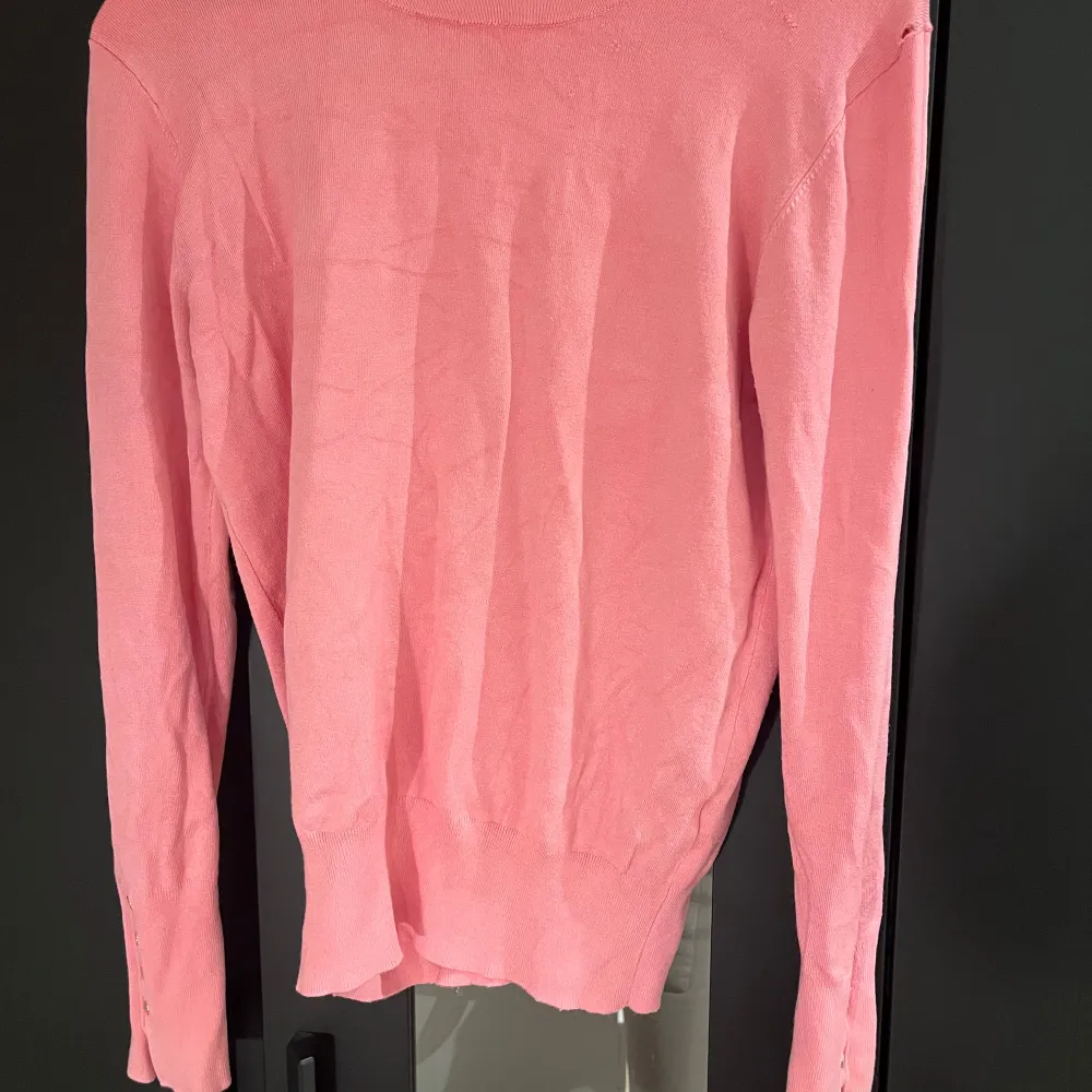Säljer en rosa tröja från Zara med rund hals och långa ärmar. Tröjan har dekorativa knappar vid ärmsluten och är perfekt för en stilren look. Observera att det finns ett litet hål vid axeln.. Neuletakit & Villapaidat.