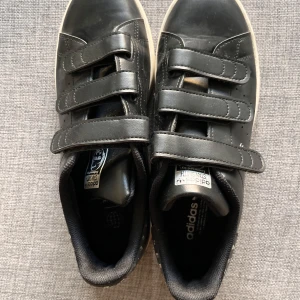 Svarta Adidas Stan Smith med  coola detaljer! - Snygga svarta Adidas Stan Smith sneakers med praktiska kardborreband och glittrande detaljer på hälen. Perfekta för en stilren look med en twist. Sulan är vit och ger en klassisk kontrast till det svarta lädret. Som nya!