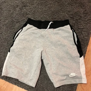 Grå shorts från Nike - Säljer ett par grå Nike shorts med svart resårmidja och vit logga. Shortsen har snörning i midjan och är perfekta för träning eller avslappnad stil. De är tillverkade i mjukt material för extra komfort. Har en fläck se bild 