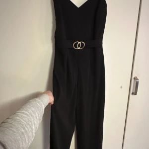 Svart ärmlös jumpsuit med bälte - Elegant svart ärmlös jumpsuit med v-ringning och ett stilrent bälte med gulddetaljer i midjan. Sparsamt använd.