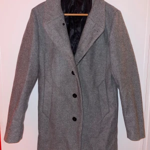 Grå kappa med knappar (Jack & Jones) - Elegant grå kappa med knappar framtill och klassisk krage. Perfekt för kyligare dagar med sin stilrena design och långa ärmar. Passar bra till både vardag och mer formella tillfällen.