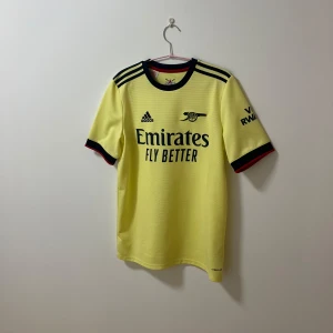 Arsenal fotbollströja - Säljer en gul fotbollströja från Adidas med korta ärmar och svarta detaljer. Tröjan har tryck med 'Emirates Fly Better' och klubbmärke på bröstet. Perfekt för fotbollsfans!