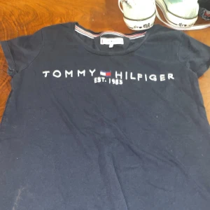 Mörkblå t-shirt från Tommy Hilfiger - Snygg mörkblå t-shirt från Tommy Hilfiger med klassisk logga och text 'EST. 1985' på framsidan. Perfekt för en avslappnad stil. Tillverkad i mjukt material för extra komfort.