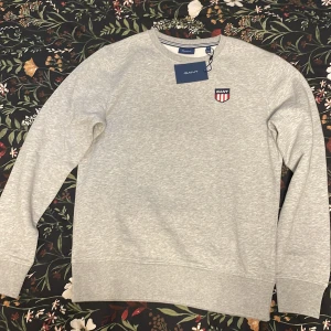 Grå sweatshirt från GANT - Säljer en stilren grå sweatshirt från GANT med rund hals och ett broderat märke på bröstet. Perfekt för en avslappnad look. Tröjan är aldrig använd och har långa ärmar och är i mjukt material. Säljes då det var fel storlek. Fick den i julklapp. Vid frågor eller pris diskution är det bara att höra av sig!