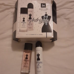 Jean Marc Intrigue Eau de Toilette och Body Spray - Set med Jean Marc Intrigue Eau de Toilette (50 ml) och Body Spray (75 ml). Flaskan har en elegant design med en svart siluett av en klänning på framsidan. Förpackningen är stilren med svart och vit färgskala. Helt nya bara testade