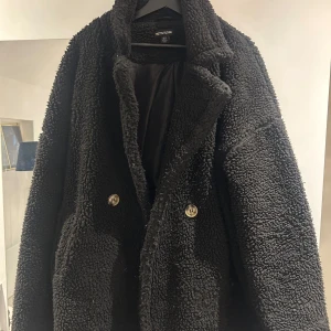 Svart teddyjacka från PLT - Mysig svart teddyjacka från Prettylittlething med knappar framtill. Perfekt för kyliga dagar med sin fluffiga och varma design. Jackan har en avslappnad passform och är långärmad. Normal i storleken. Nypris 800kr