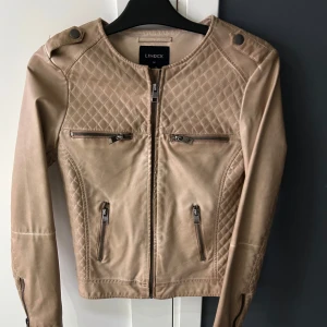 Beige jacka från Lindex - Snygg beige jacka från Lindex med quiltade detaljer och dragkedjor. Jackan har en rund hals och långa ärmar med dragkedjor vid ärmsluten. Perfekt för en stilren look.